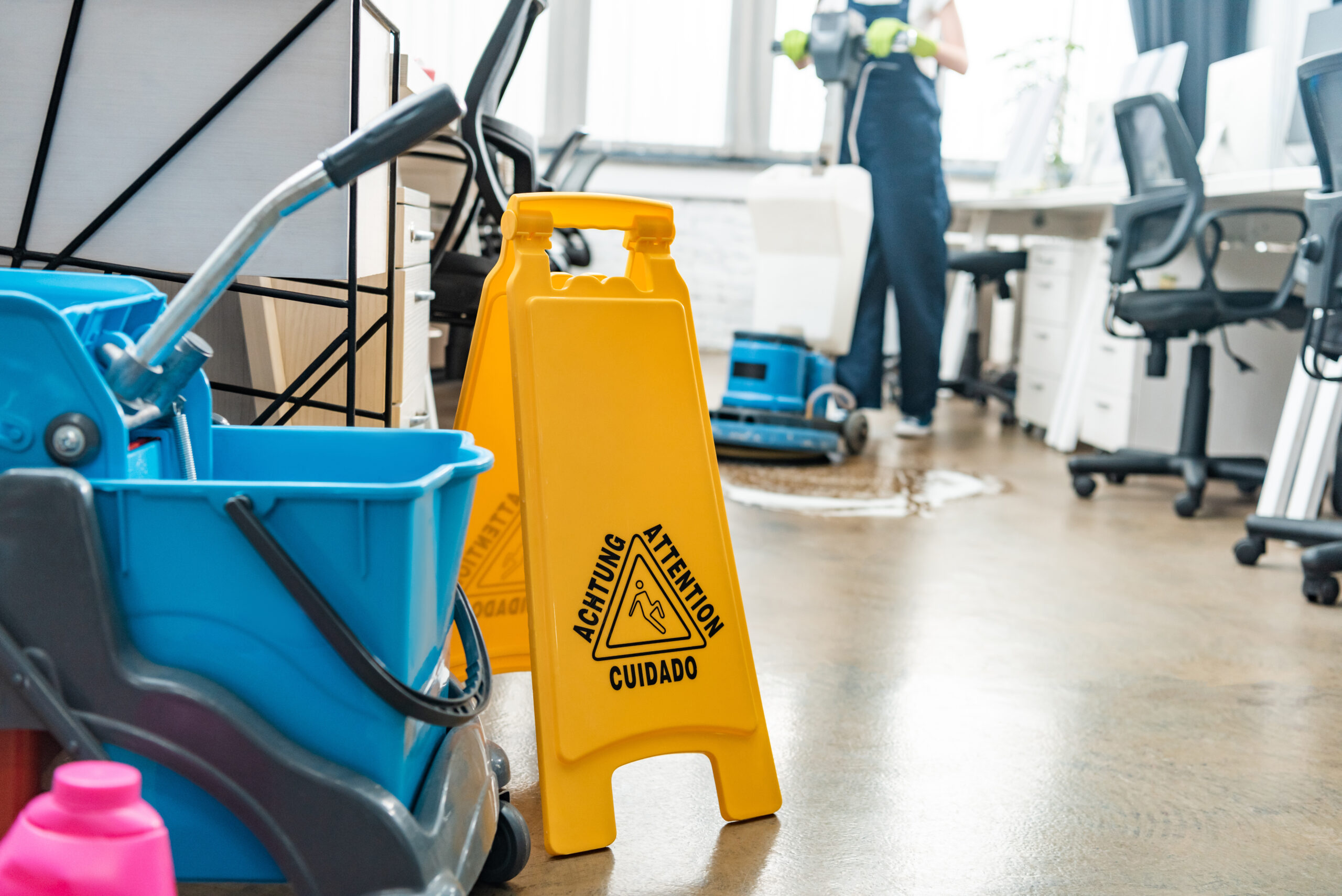 selective focus of cart with buckets and wet floor caution sign, and cleaner washing floor with selective focus of cart with buckets and wet floor caution sign, and cleaner washing floor with